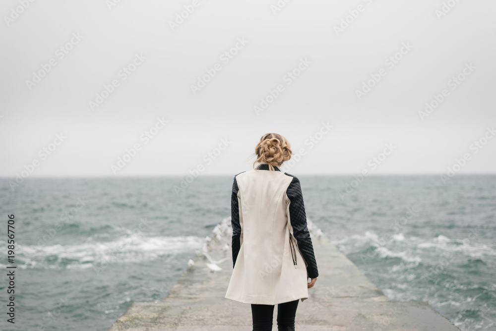 The girl stands with her back on the pier and looks at the sea.