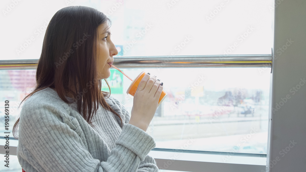 Fototapeta premium Young woman drinks freshly squeezed juice in a cafe, looking out the window