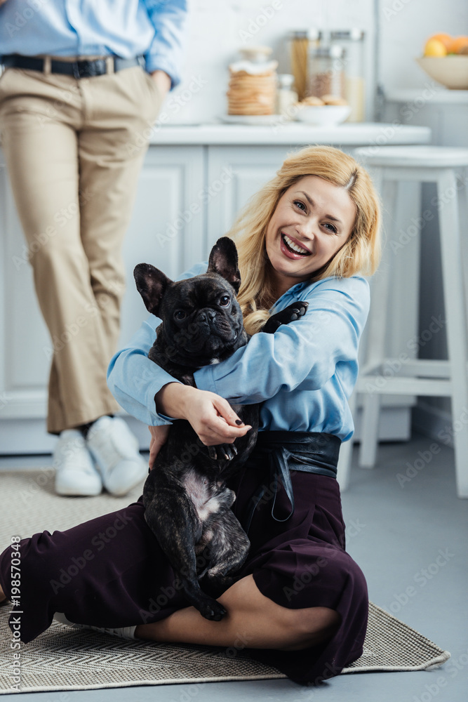 Man standing by happy woman hugging french bulldog on kitchen floor Man standing by happy woman hugging french bulldog on kitchen floor