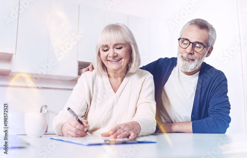 Place for signature. Cheerful delighted old couple sitting in the kitchen and signing the insurance contract while expressing positivity