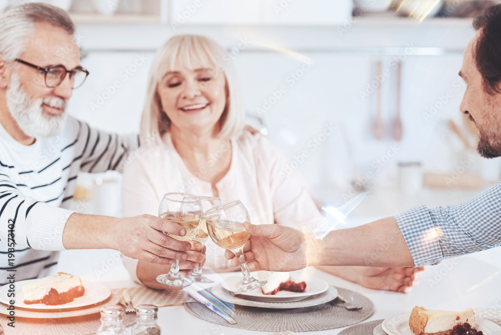 Enjoyable time together. Positive smiling loving aged parents drinking wine with their son while sitting in the kitchen and having family celebration Enjoyable time together. Positive smiling loving aged parents drinking wine with their son while sitting in the kitchen and having family celebration