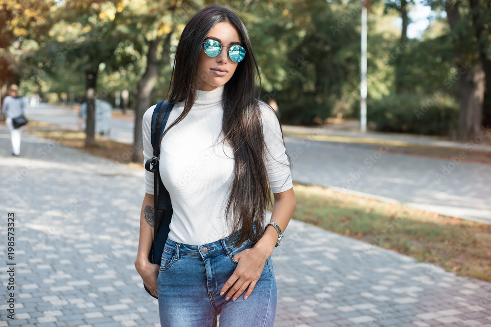 Fototapeta premium Hipster girl wearing blank gray t-shirt, jeans and backpack posing in the park, minimalist urban clothing style.Stylish happy young woman wearing boyfriend jeans and long jeans coat
