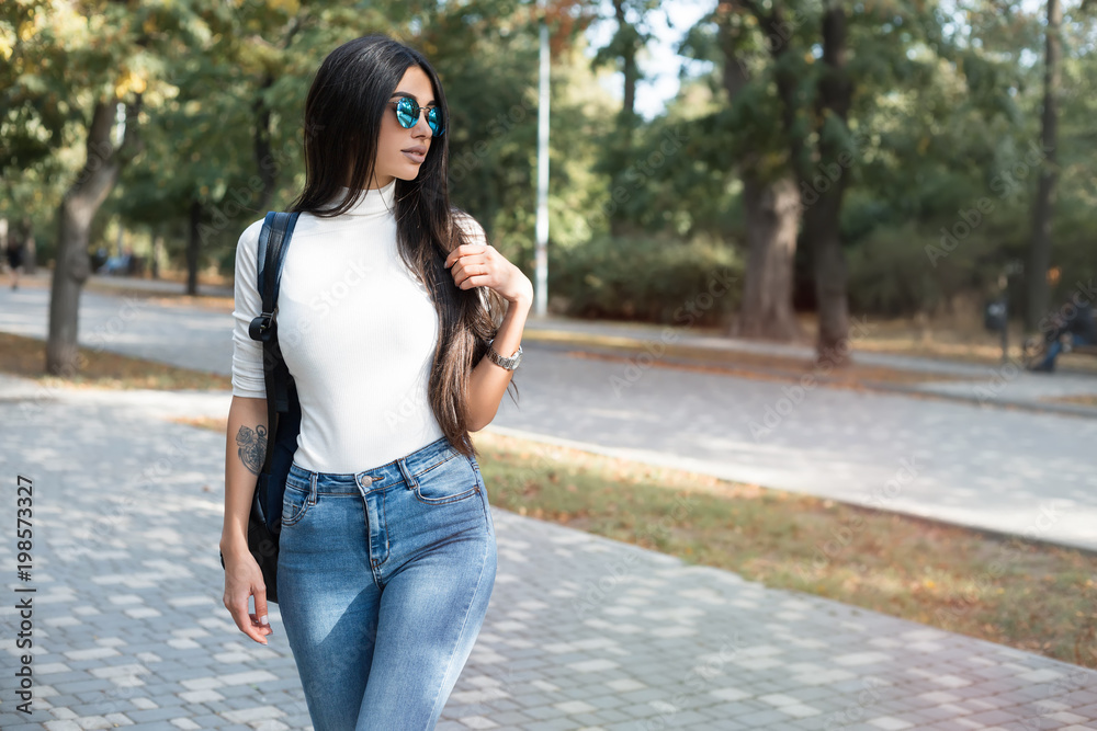 Hipster girl wearing khaki white sweat,t-shirt, jeans and back pack posing  walking in autumn city, minimalist urban clothing style.Stylish happy young  woman wearing boyfriend jeans and blue sunglasses Stock Photo | Adobe