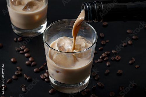Pouring irish cream in a glass with ice, surrounded by coffee beans on a dark black background photo