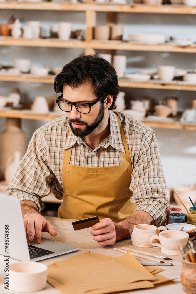 Fotografie bearded man shopping online with laptop and credit card in pottery workshop