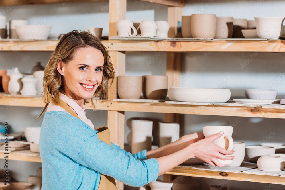 beautiful smiling potter with ceramic dishware on shelves in workshop