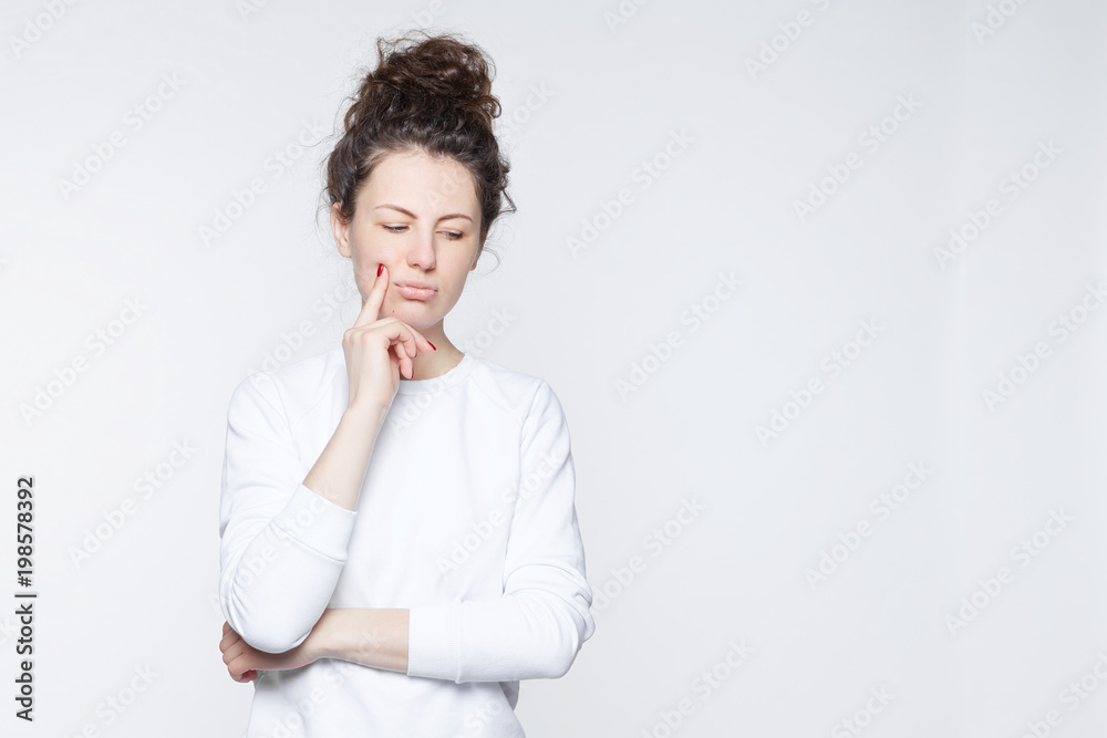 Young woman with hair bun, blue eyes frowning eyebrows, looking down, holding index finger on chin having doubt and suspicion feeling sceptic about something. Human emotions and expressions concept. Young woman with hair bun, blue eyes frowning eyebrows, looking down, holding index finger on chin having doubt and suspicion feeling sceptic about something. Human emotions and expressions concept.