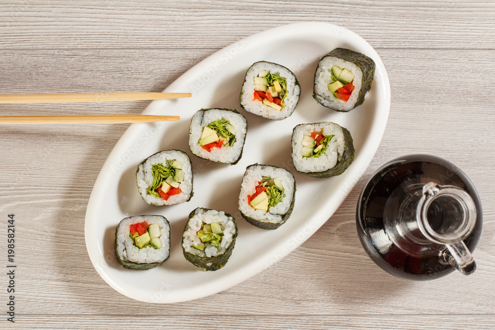 Sushi rolls with rice, pieces of avocado, cucumber, red bell pepper and lettuce leaves on ceramic plate, glass bottle with soy sauce Sushi rolls with rice, pieces of avocado, cucumber, red bell pepper and lettuce leaves on ceramic plate, glass bottle with soy sauce