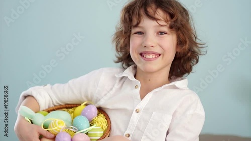 Curly red-haired child in a white shirt with a figure of an Easter bunny and a set of Easter eggs in a basket. A boy laughs cheerfully. Slow Motion. photo