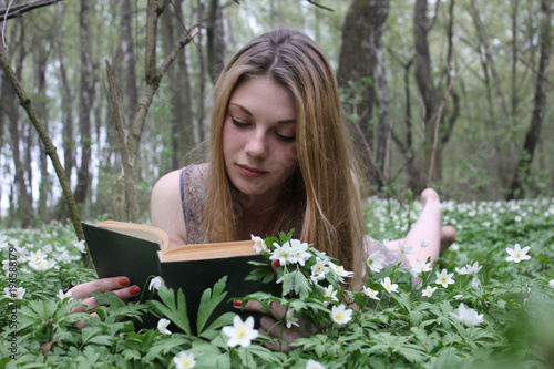 Young teen girl reading a book. Luxurious long hair. Blonde girl in summer dress. Woman resting lying on the grass. Meadow of white flowers in the forest. Warm spring day. Photo shoot outdoors.