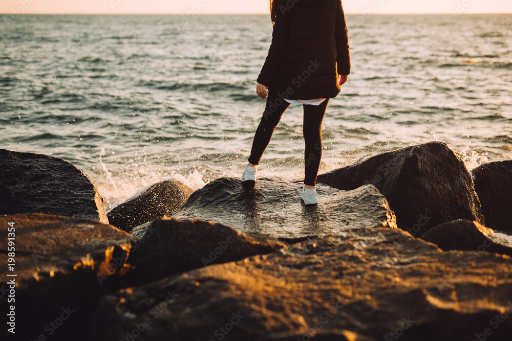 Young girl stands on the stones near the sea. From behind it is beautiful sunset