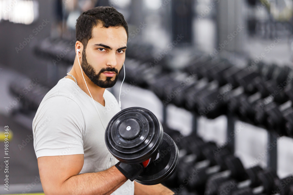 Strong sexy bearded man wears white t-shirt and headphones does arm biceps  workout with dumbbells in the gym Photos | Adobe Stock