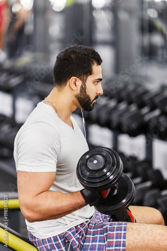 Handsome sexy bearded man wears white t-shirt and headphones does arm biceps workout with dumbbells in the gym