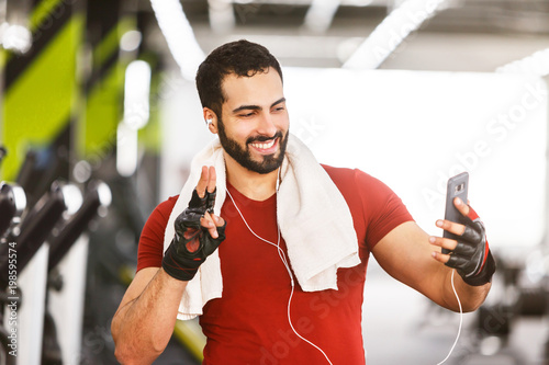 Bearded muscular smiling man using smartphone for videochat in the gym, hardworking rest photo