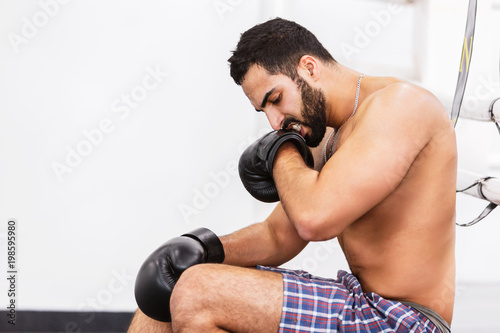Muscular handsome topless boxer wears black gloves sitting near the ring