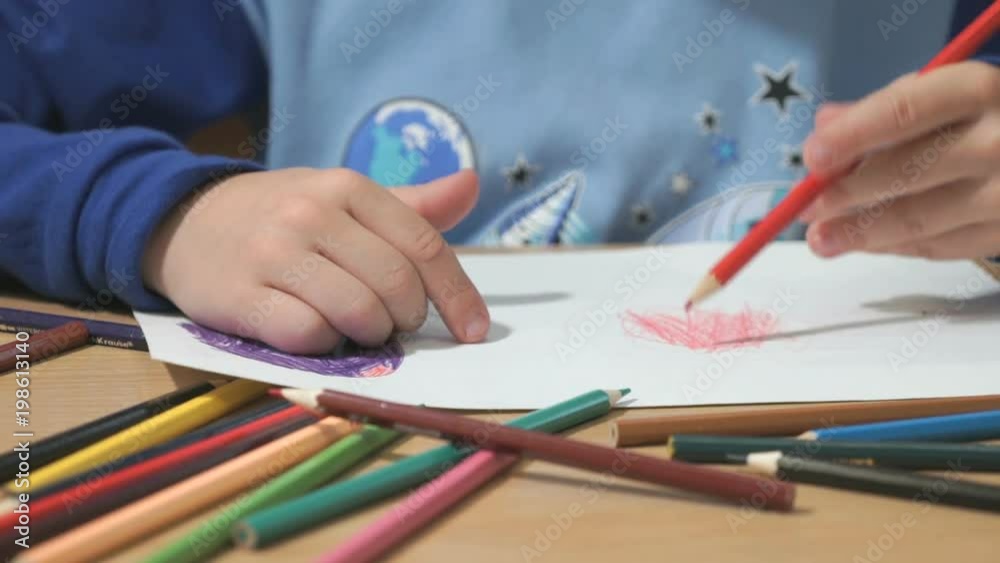 Close-up of hands of unidentified little child in blue sweater sitting at table drawing pictures using multicolored pencils in kindergarten Close-up of hands of unidentified little child in blue sweater sitting at table drawing pictures using multicolored pencils in kindergarten