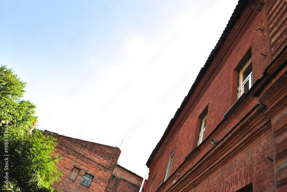 Old red brick building walls with windows, green trees, view from ground on slate roof,  blue sky background