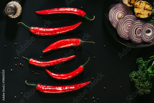 top view of red chili peppers, peppercorns, sliced onion and grilled garlic on black