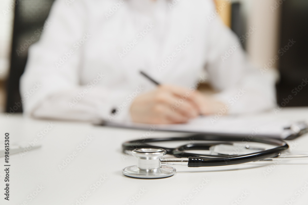 Female doctor hands working filling out medical documents in light office in hospital. Woman writing on paper in clipboard, focus on stethoscope on table in consulting room. Medicine concept. Top view