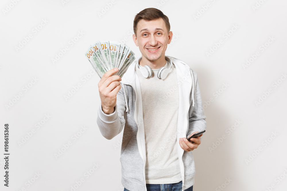 Young handsome smiling man student in t-shirt, light sweatshirt with headphones around neck holds bundle of dollars, cash money, mobile phone in hands isolated on white background. Concept of success. Young handsome smiling man student in t-shirt, light sweatshirt with headphones around neck holds bundle of dollars, cash money, mobile phone in hands isolated on white background. Concept of success.