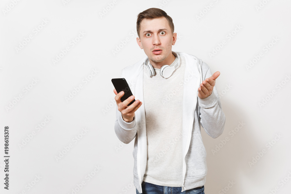 Young concerned handsome man student in t-shirt, light sweatshirt with headphones around neck is surprised with mobile phone in hands isolated on white background. Concept of communication, emotions. Young concerned handsome man student in t-shirt, light sweatshirt with headphones around neck is surprised with mobile phone in hands isolated on white background. Concept of communication, emotions.