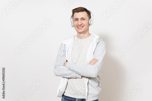 Young handsome smiling man student in t-shirt and light sweatshirt listening to music with white wireless headphones and crossed arms over chest isolated on white background. Concept of emotions © ViDi Studio