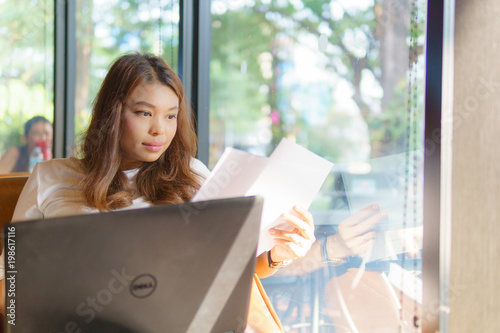 beautiful asian young business woman sitting near transparent window and holding business document with laptop for her job at cafe or coffee shop. business success concept.