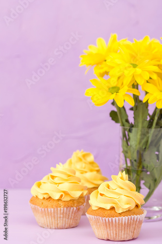 Yellow chrysanthemum in glass and cupcake with yellow cream on violet pastel background. Mother's day layout design with flowers and dessert.