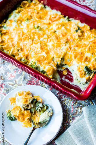 Overhead shot of browned chicken divan casserole in red baking dish on gray floral linen with individual serving on white plate with silver spoon.