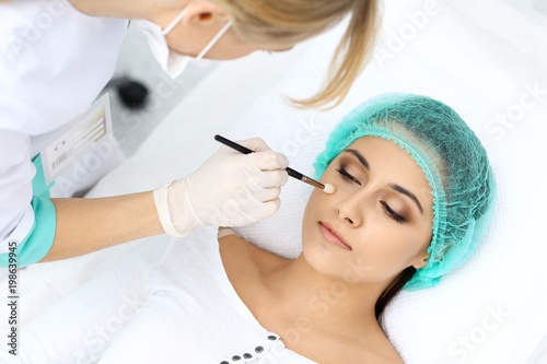 Professional beautician is doing cosmetic procedure at light medical background touching patient's face with brush, closeup. Cosmetology treatment