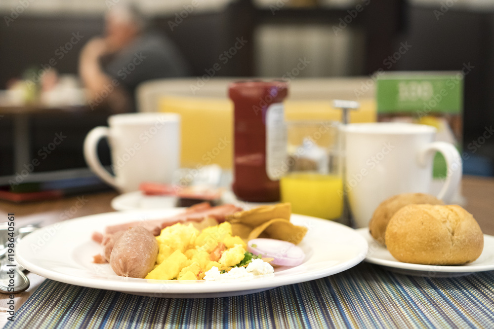 Breakfast on table served with egg, bread, thickness ham, orange juice, coffee in white ceramic cup, with blurry background.