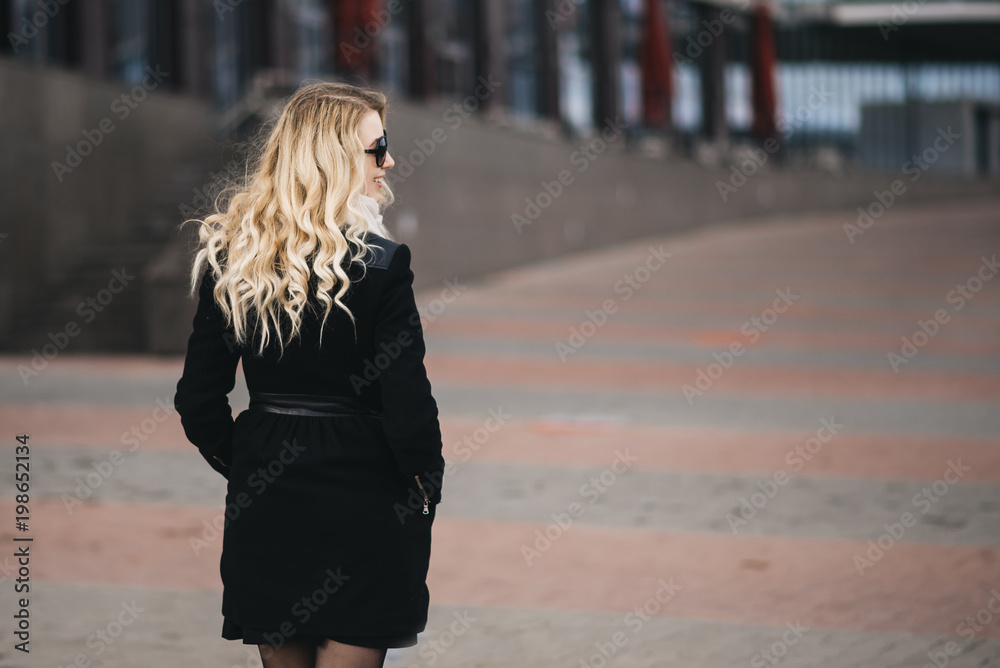 Beautiful young girl with blond wavy hair in a black coat against the backdrop of modern buildings Beautiful young girl with blond wavy hair in a black coat against the backdrop of modern buildings