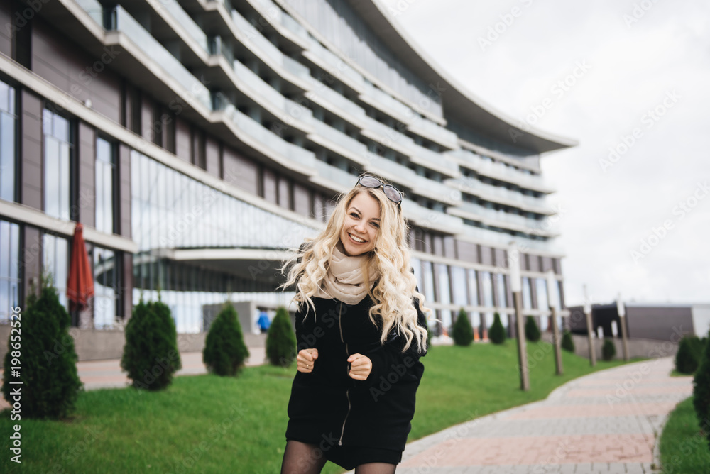 Beautiful young girl with blond wavy hair in a black coat fun on background of modern building Beautiful young girl with blond wavy hair in a black coat fun on background of modern building