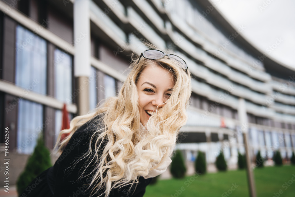 Beautiful young girl with blond wavy hair in a black coat fun on background of modern building