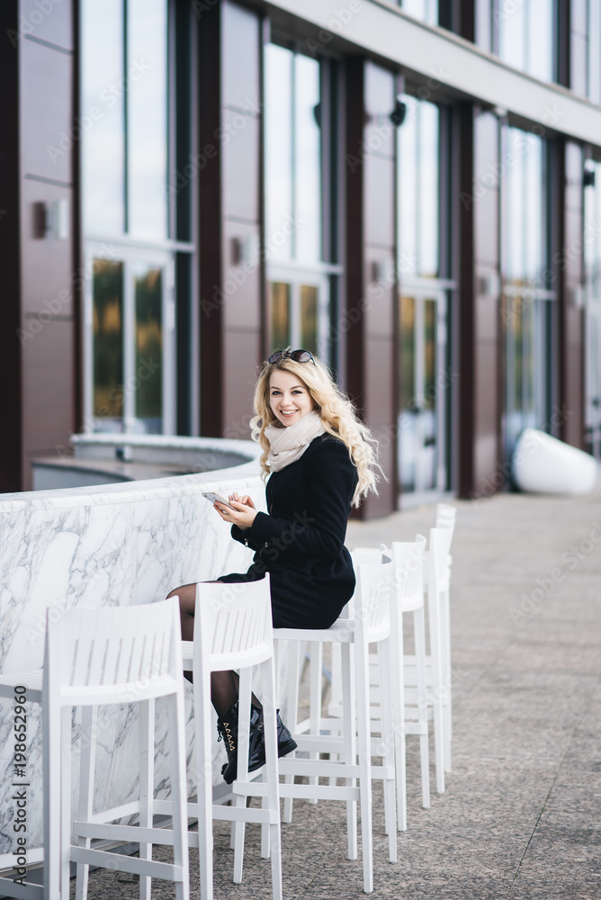 Beautiful attractive blonde girl sitting behind a White marble bar counter with high chairs on the background of a modern building in minimal style