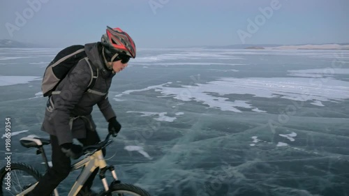 Man is riding a bicycle on ice. The cyclist is dressed in a gray down jacket, backpack and helmet. Ice of the frozen Lake Baikal. The tires on the bicycle are covered with special spikes. The traveler photo