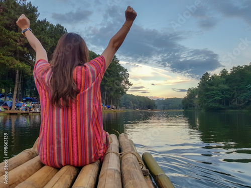 back view of asian female sitting on bamboo boat looking to amazing view sunset over the river among the jungle  and raise her arm for happiness and fresh air.