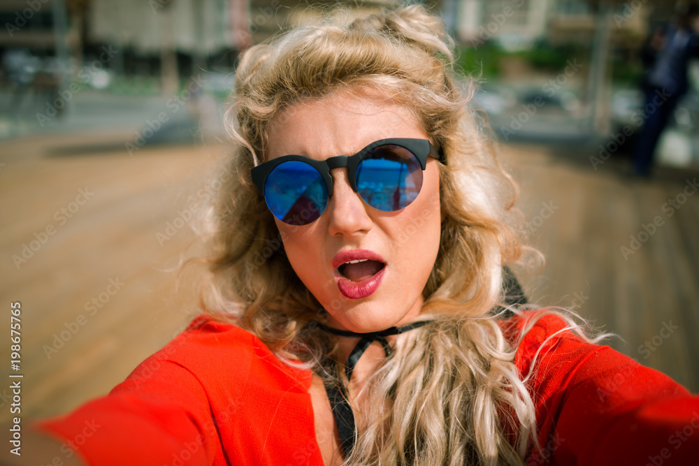 Surprised elegant blonde girl on background of wooden promenade. Close-up view of young pretty woman in bright red dress with beautiful face at the city waterfront