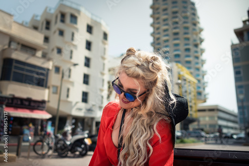 Young attractive woman in bright red dress sits on on the bench on the background of cityscape. Elegant blonde girl with pretty smile on her face is enjoying warm summer day