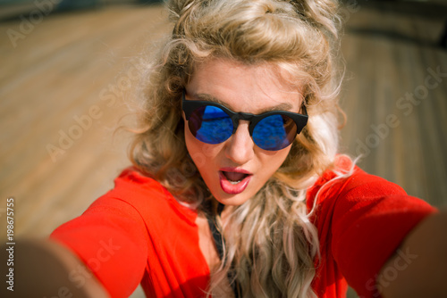 Close-up view of young sensual woman in bright red dress with surprise on her face at the city waterfront. Elegant blonde girl make a selfie on background of wooden promenade
