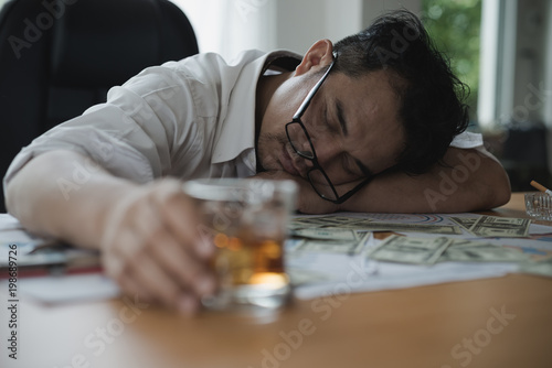 Stressed asian businessman holding a glass of whiskey. he sleep on the money, data charts, business document at office desk. alcohol addiction - drunk businessman concept