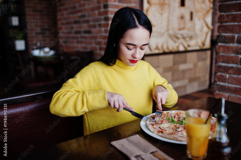 Funny brunette girl in yellow sweater eating pizza at restaurant.