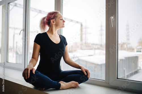 Sensual young girl with pink hair is meditating while sitting in lotus position near window. Her eyes are closed with peace and pleasure. Free Download Sensual young girl with pink hair is meditating while sitting in lotus position near window. Her eyes are closed with peace and pleasure. - GFXLoad