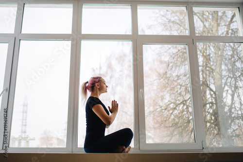 Sensual young girl with pink hair is meditating while sitting in lotus position near window. Woman holds her hands in Namaste posture, do yoga lotus position. side view