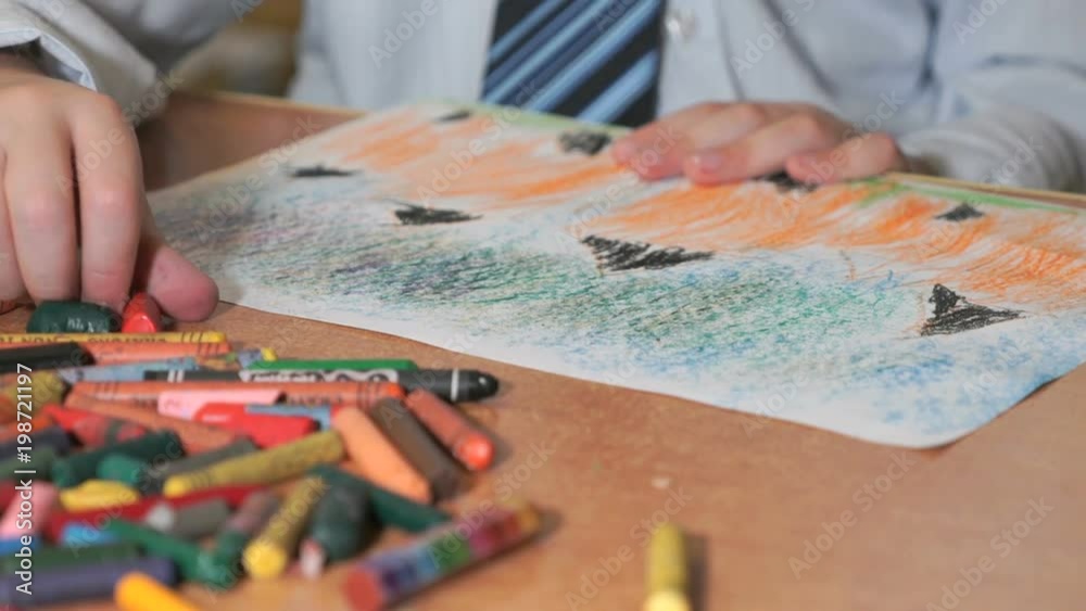 Close-up of unknown little boy dressed in shirt with tie draws the pictures using color chalks and pencils in the album in the kindergarten