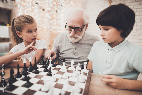 Grandfather, grandson and granddaughter at home. Grandpa teaches children how to play chess.