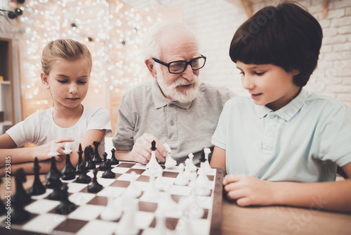 Grandfather, grandson and granddaughter at home. Children and grandpa are playing chess.