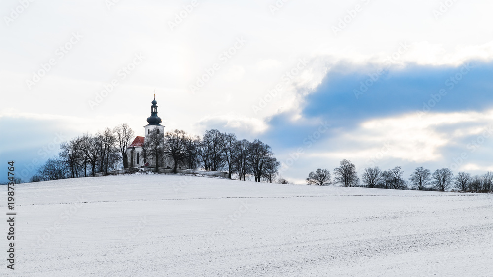 Church in idyllic winter landscape. Kostelec, South Bohemia, Europe. Artistic scene with chapel on hill, snowy field and blue sky with white clouds. It symbolizes religion, Christianity, Christmas. Church in idyllic winter landscape. Kostelec, South Bohemia, Europe. Artistic scene with chapel on hill, snowy field and blue sky with white clouds. It symbolizes religion, Christianity, Christmas.