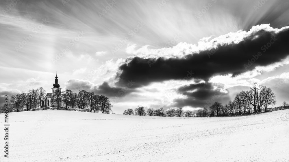 Artistic black and white landscape with a church on hillock. Kostelec, South Bohemia, Europe. Wintry scene with a snowy field, churchyard with chapel and a dramatic sky with clouds and sunbeams.