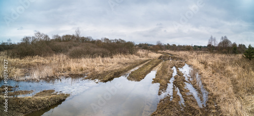 Fototapeta Naklejka Na Ścianę i Meble -  Off-road track with tire imprints and stagnant water in winter landscape. Spring thawing in deserted military area with the muddy bumpy path and a blue sky.